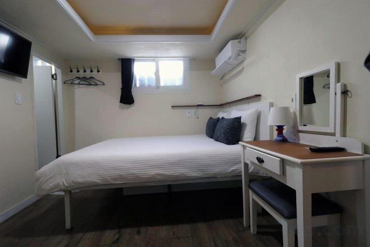 A bedroom with a bed, a desk, a chair, a TV, and a window.