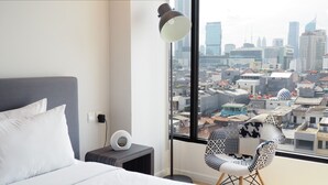 A hotel room with a bed, a bedside table with a lamp, a chair, and a large window offering a city view. - Juno Tanah Abang Jakarta (Jakarta)