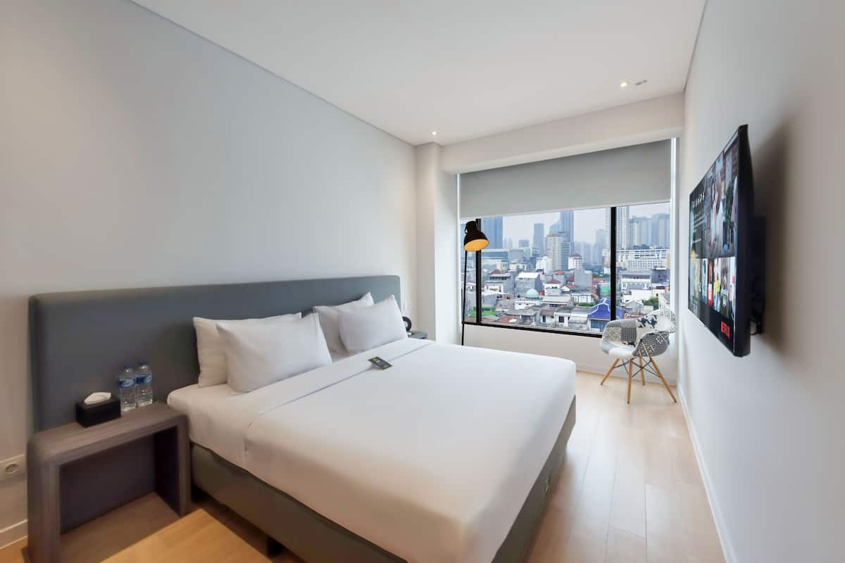Een moderne hotelkamer met een groot bed, een nachtkastje met een lamp, een stoel en uitzicht op de stad door grote ramen.