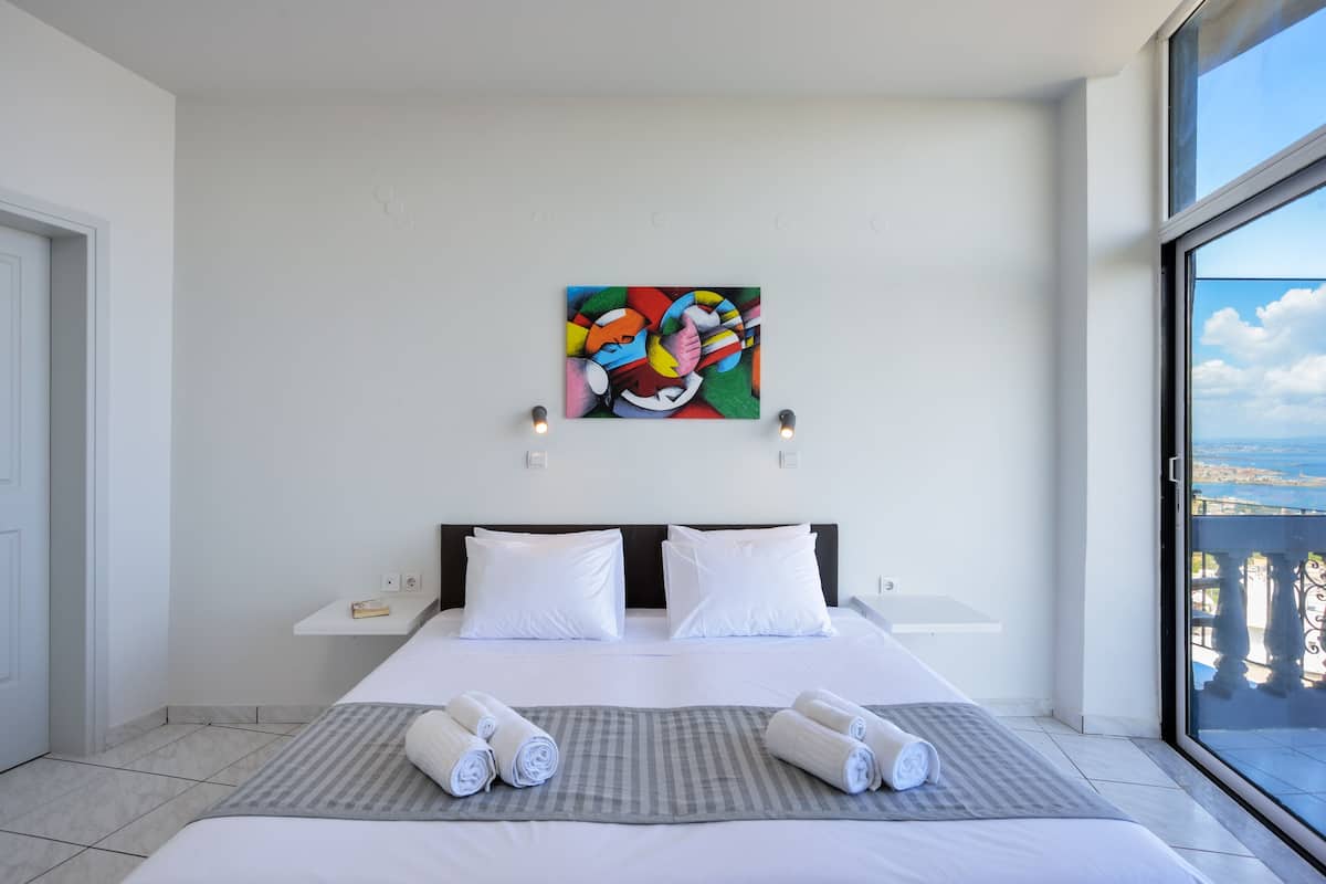 A modern hotel room with a large bed, two white pillows, a colorful abstract painting on the wall, and a view of the sea through a large window.