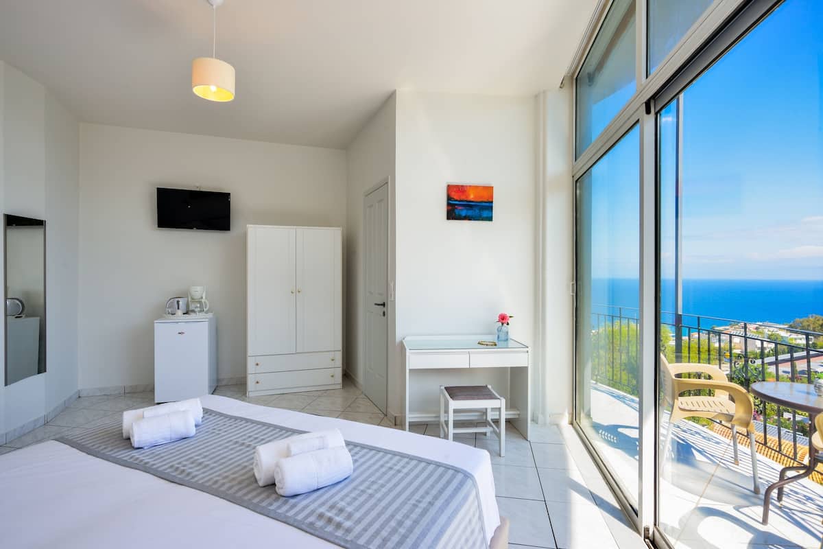 A hotel room with a bed, a desk, a TV, a minibar, and a balcony with a view of the sea.