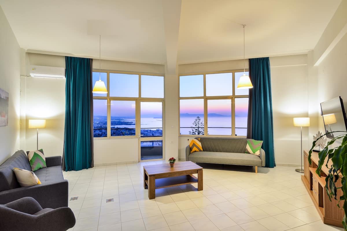 A spacious living room with a sofa, a coffee table, and a TV. Large windows offer a view of the cityscape.