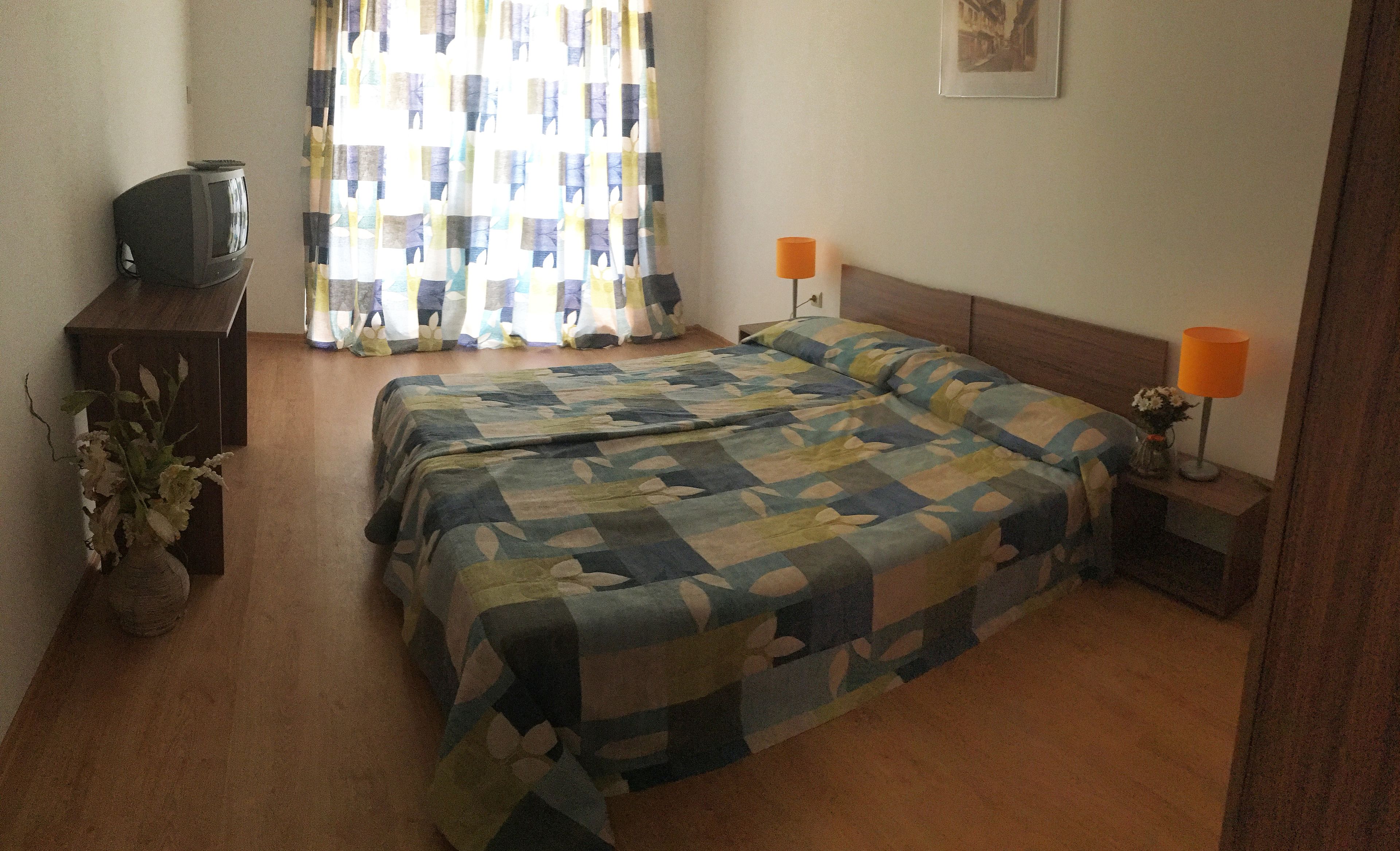 A bedroom with two beds, a wooden nightstand with a lamp, a small table with a lamp, and a television on a stand.