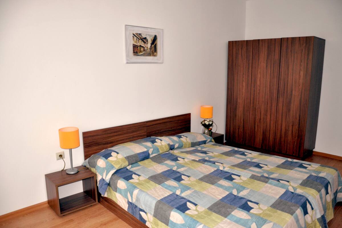 Een slaapkamer met een houten hoofdbord, een gewatteerde sprei, twee bedlampen en een lijst met een afbeelding aan de muur.