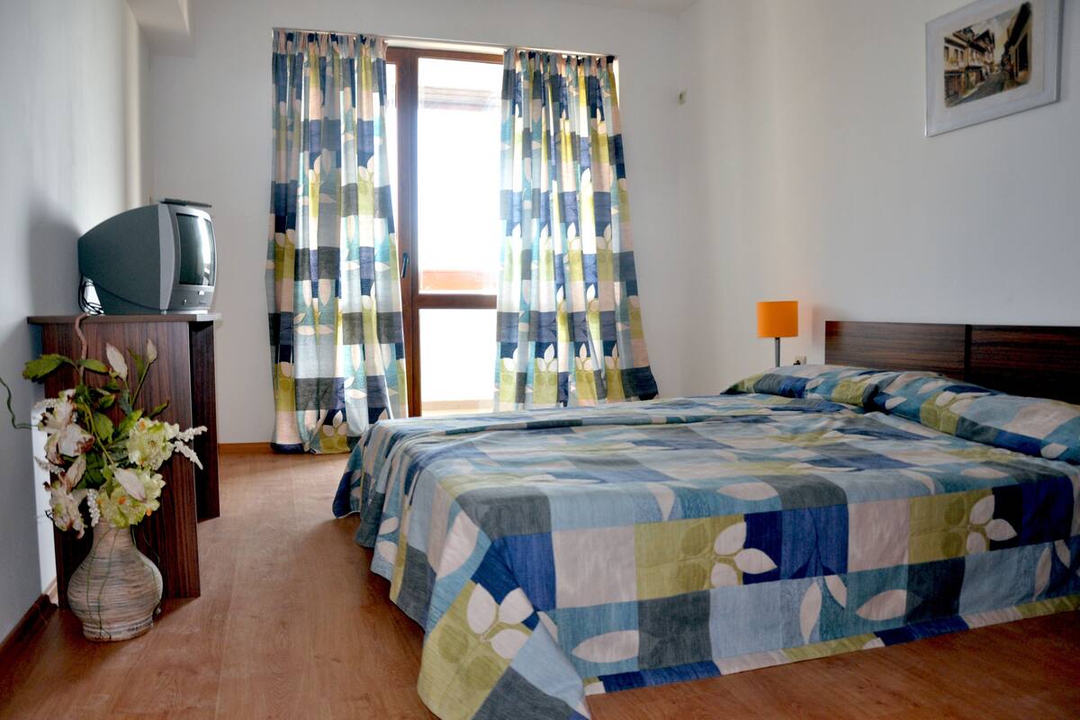 Een slaapkamer met een bed, een televisie op een houten dressoir en een vaas met bloemen.