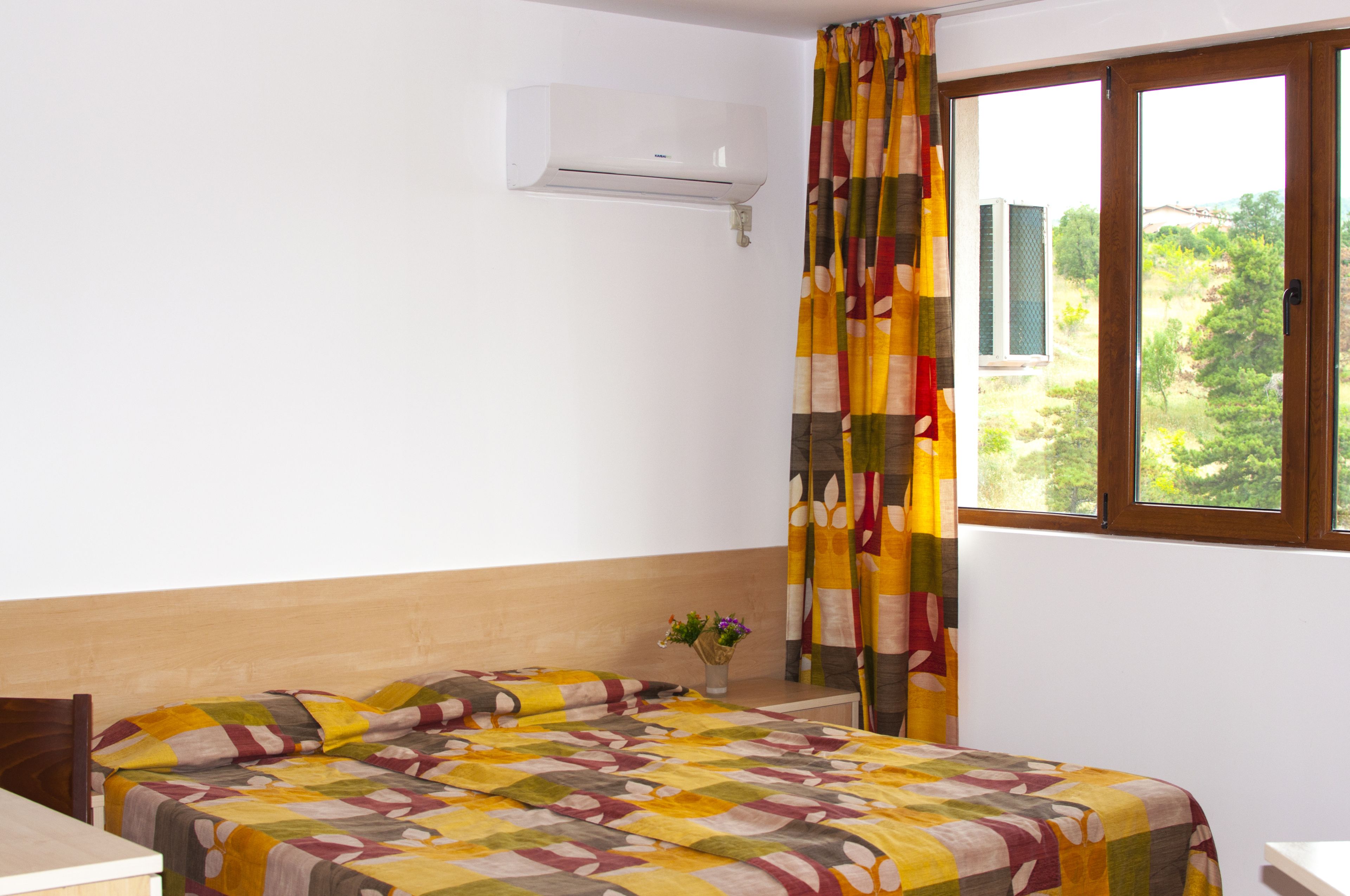 A bedroom with a bed, a window with curtains, a wall-mounted air conditioner, and a small table with a vase of flowers.