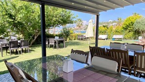 An outdoor dining area with glass tables and chairs, a covered seating area, and a view of the marina and greenery. - Cilek Butik Otel (Cesme)