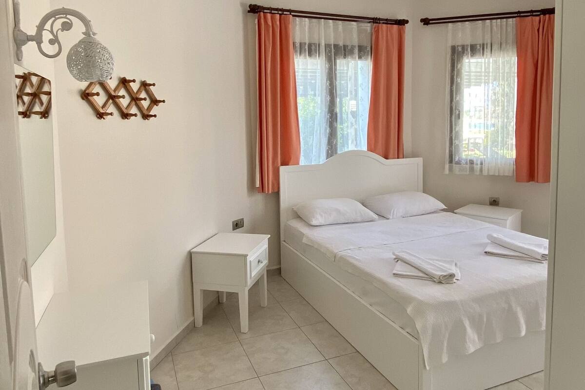A bedroom with a bed, a nightstand, a mirror, and a window with curtains.