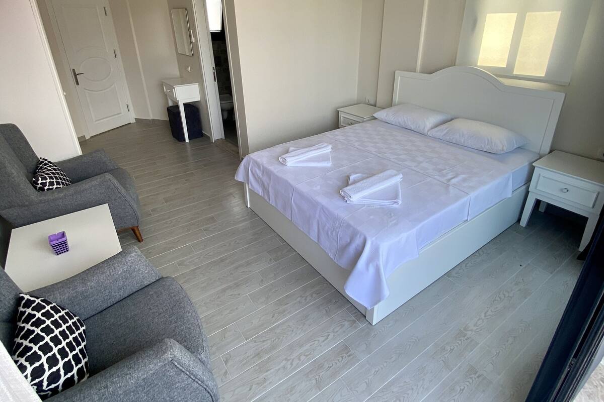 A bedroom with a bed, a grey sofa, a white bedside table, a white wardrobe, and a ceiling light fixture.