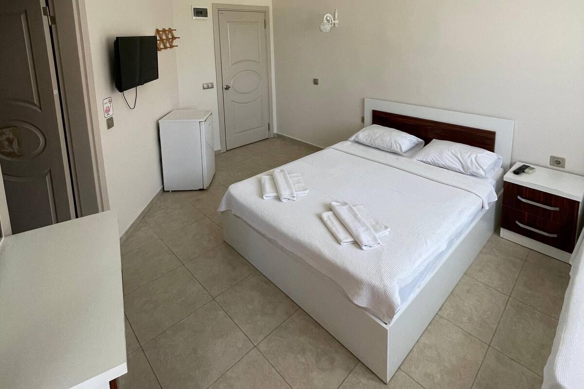 A hotel room with a bed, a bedside table, a TV mounted on the wall, a door, and a small refrigerator.