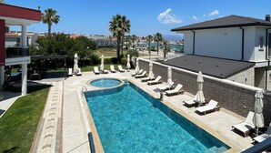 A swimming pool with lounge chairs and umbrellas, surrounded by palm trees and buildings. - Cilek Butik Otel (Cesme)