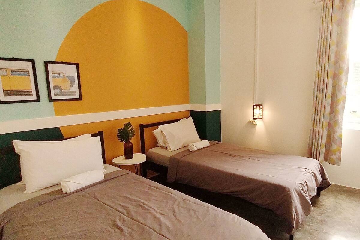 Habitación de hotel con dos camas, una mesita redonda pequeña, una lámpara de pared y un ventanal con cortinas.