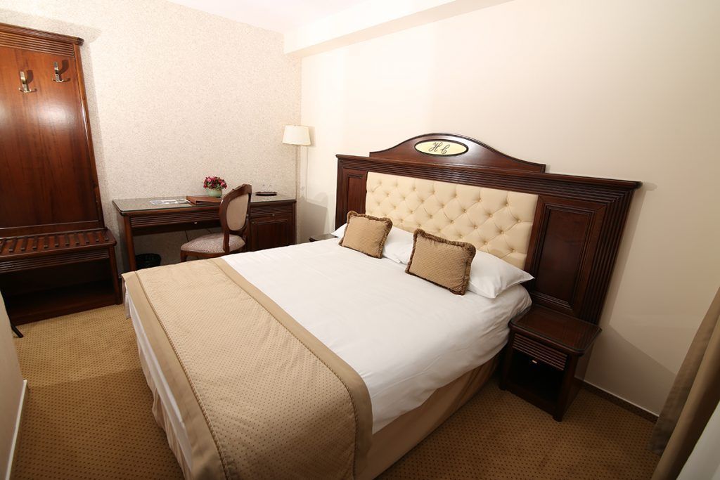 Een hotelkamer met een groot bed, houten hoofdeinde, twee nachtkastjes, een bureau met stoel, en een kledingkast.