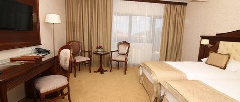 Habitación de hotel con cama, escritorio con silla, una mesita con sillas, televisor y ventana con cortinas.