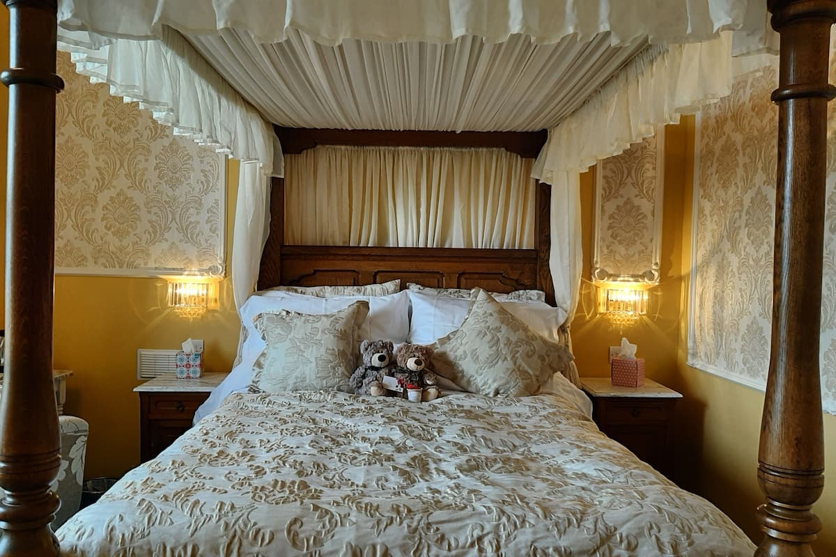 A four-poster canopy bed with a floral bedspread, two bedside lamps, and a teddy bear on the pillows.