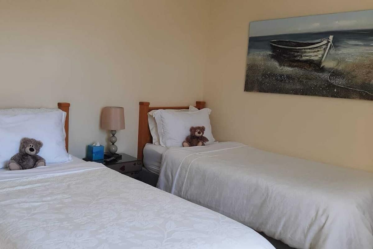 Two single beds with white linens, each with a teddy bear on the pillows. A lamp and a tissue box are on the nightstand between the beds.
