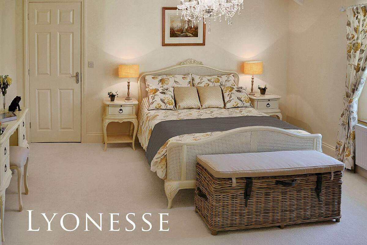 A bedroom with a bed, two bedside tables, a nightstand, a wicker chest, a chandelier, a window with curtains, and a door.