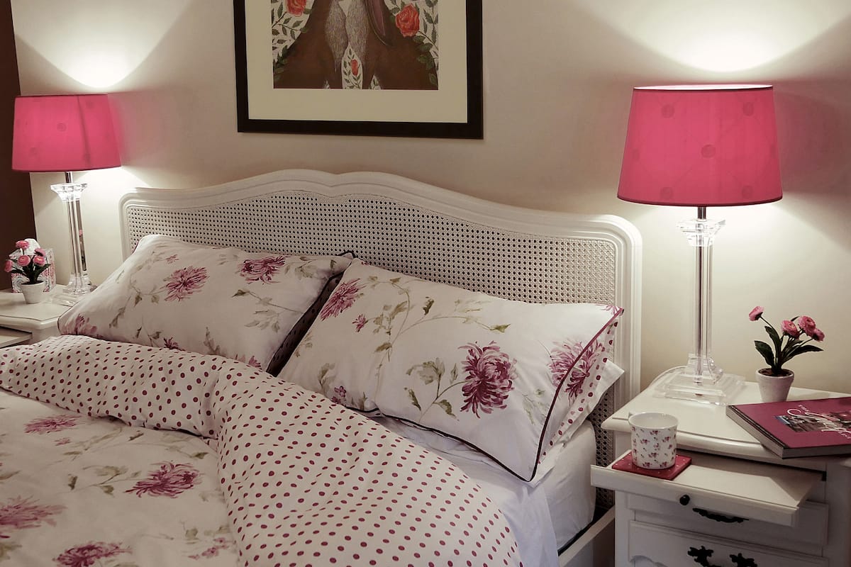 A neatly made bed with floral bedding, two bedside lamps, a framed picture on the wall, and a small table with a potted plant.