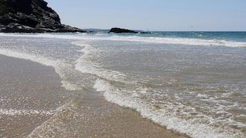 Une plage où les vagues se brisent près du rivage, une falaise rocheuse et une formation rocheuse visible au loin.