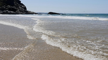 Una playa con olas que se acercan a la orilla, un acantilado rocoso y una formación rocosa distante.