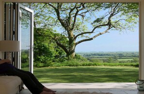 A room with a large window overlooking a grassy landscape and a tree. - Caroe Farmhouse (Camelford)