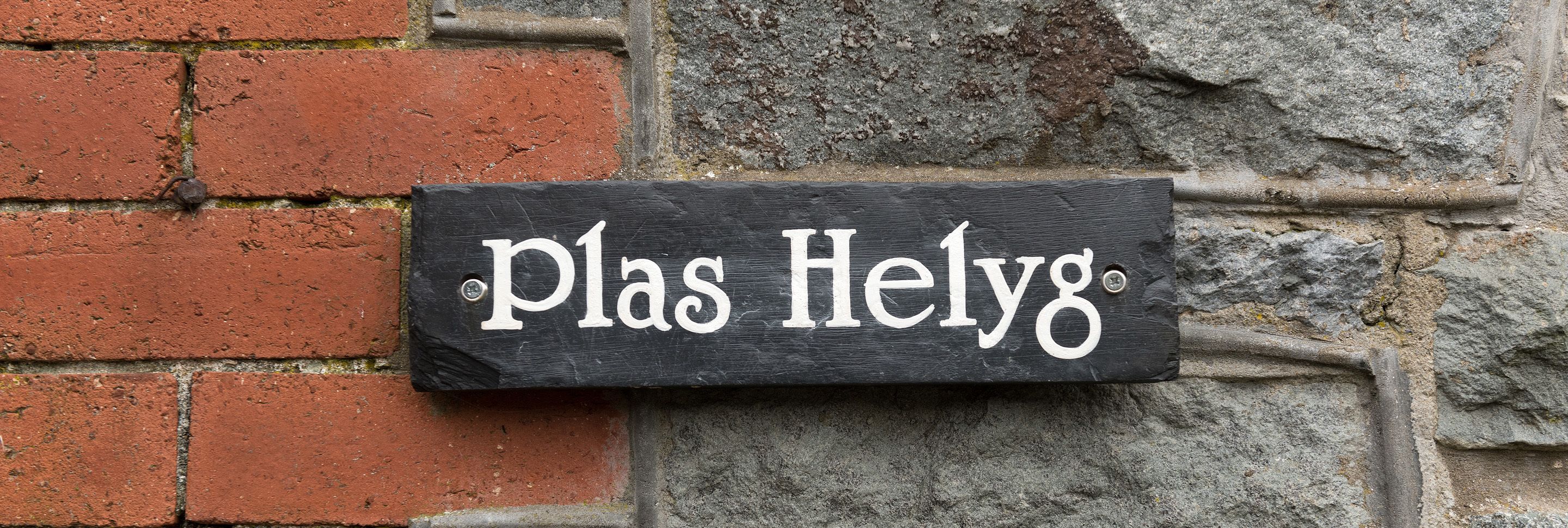 Placa de ardósia preta com letras brancas que dizem 'Plas Helyg' fixada numa parede de tijolos.