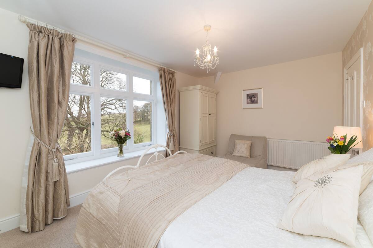 A bedroom with a large bed, a window with curtains, a radiator, a chair, and a bedside table with a lamp and flowers.