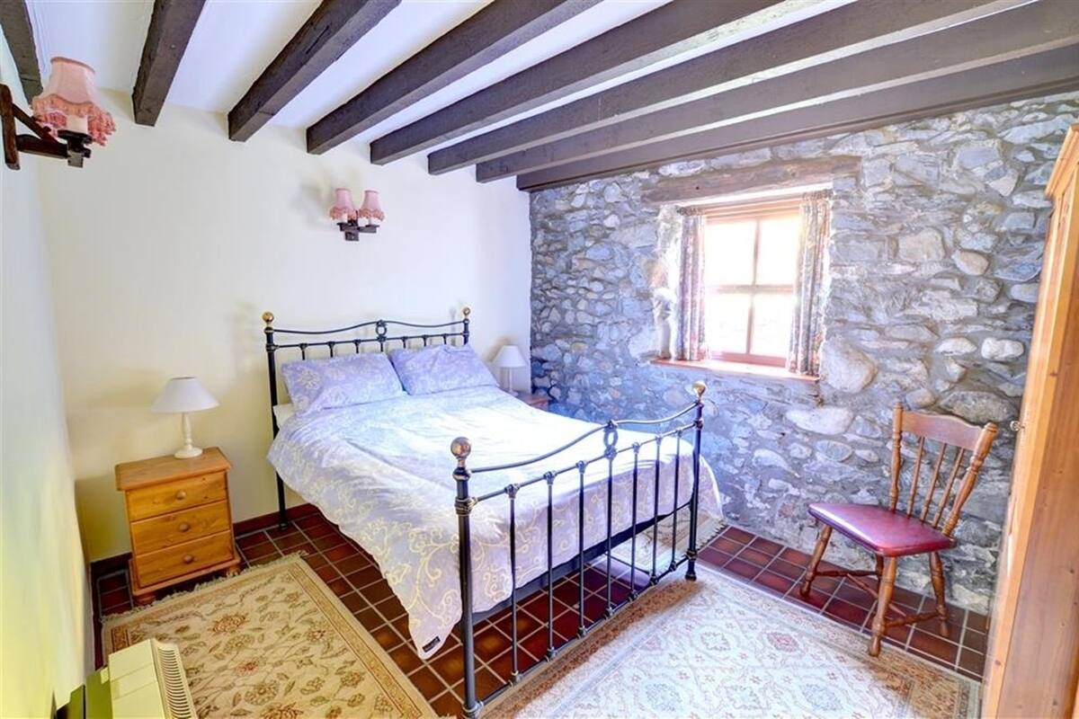 A bedroom with a stone wall, a wooden bed frame, a nightstand, a chair, and a window.