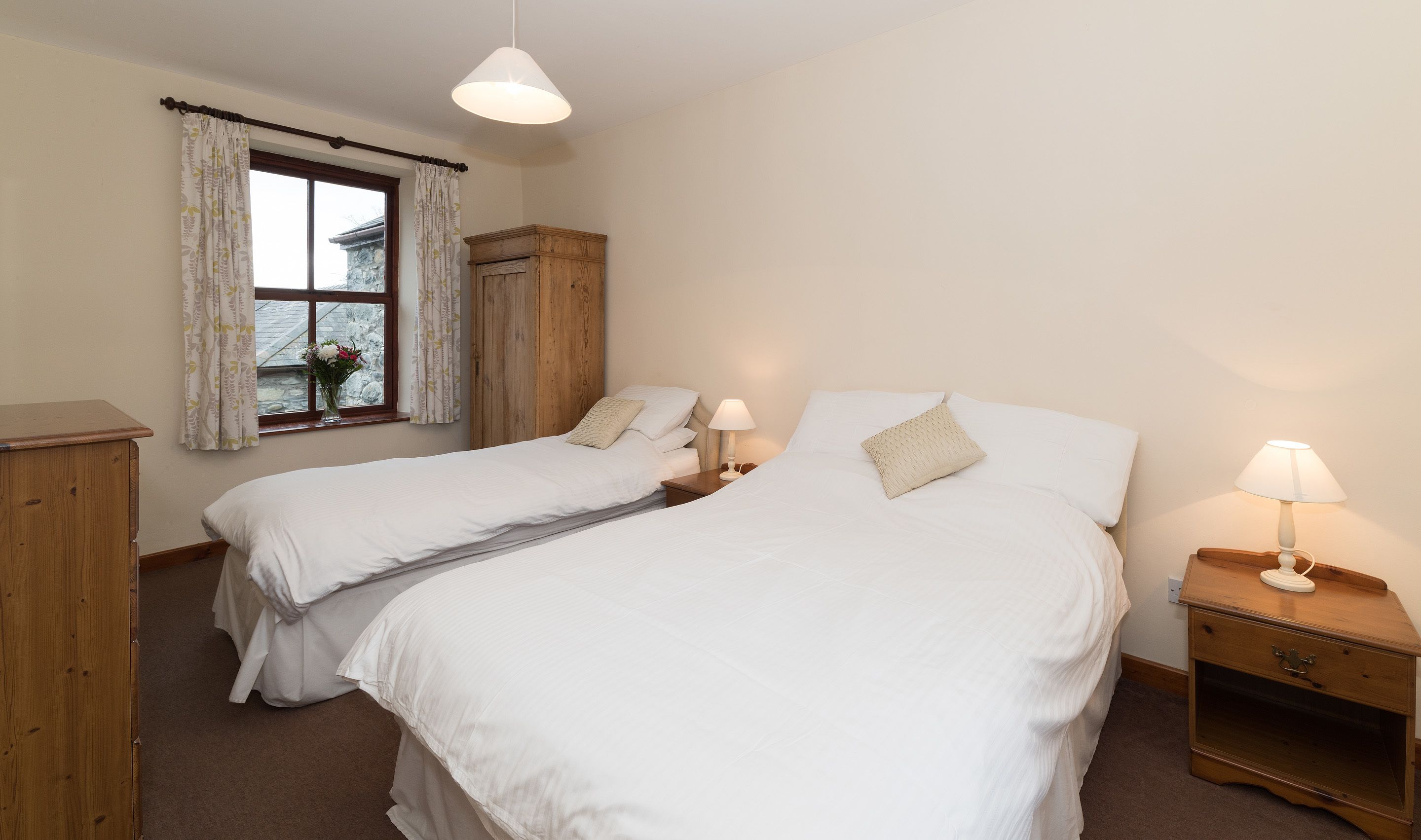 A bedroom with two single beds, a wooden wardrobe, a nightstand with a lamp, and a window with curtains.