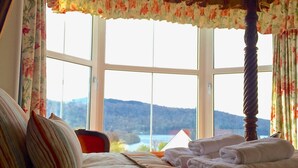 Un dormitorio con un ventanal grande, una cama con ropa de cama a rayas y vistas a un lago y colinas.