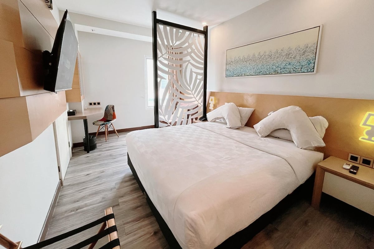 A modern hotel room with a large bed, a desk, and a chair. There is a painting on the wall and a decorative screen door.