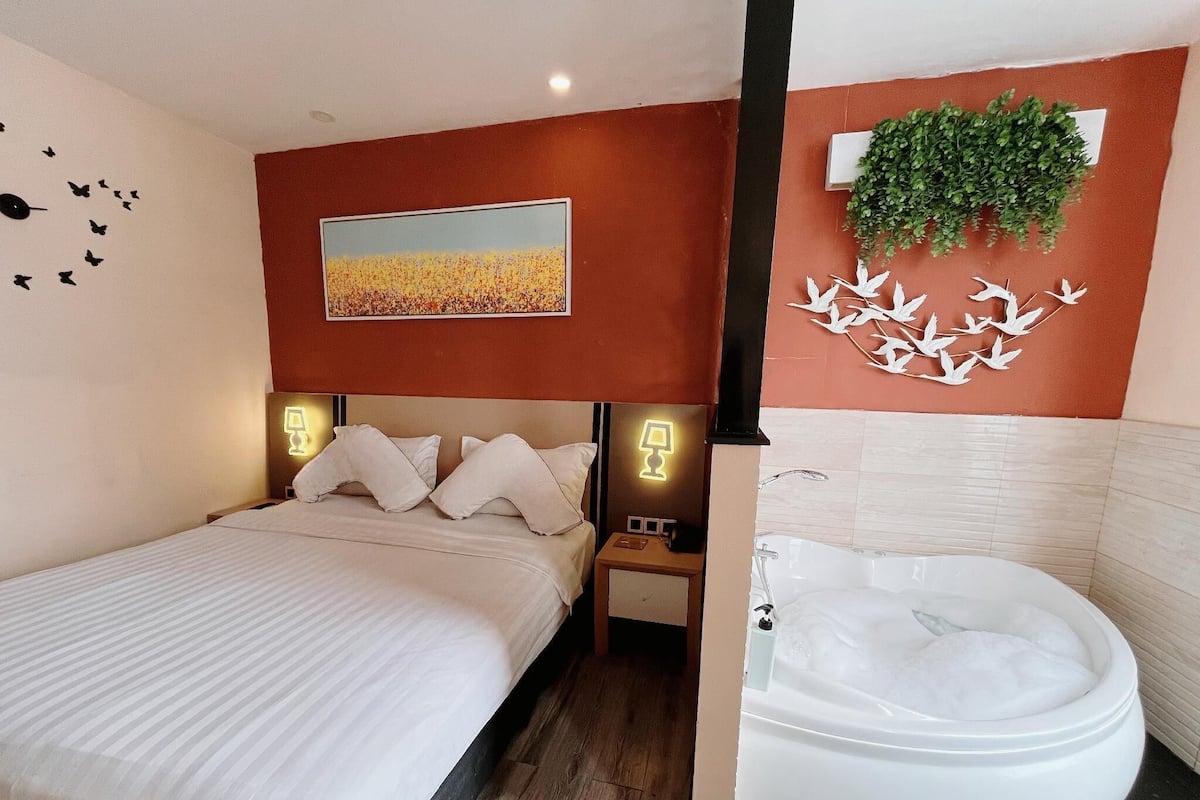 A hotel room with a bed, bedside lamps, a painting, a wall-mounted plant, and a bathroom with a bathtub.