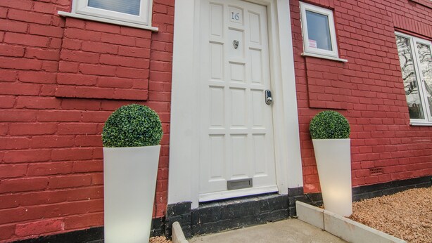 Una casa de ladrillo rojo con una puerta blanca, número 16, y dos topiarios en macetas a cada lado.