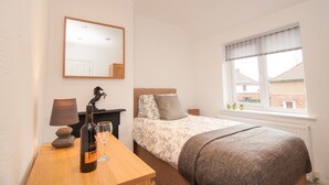 Un dormitorio con una cama, una mesita de noche con una botella y un vaso, una lámpara, un espejo y una ventana con vistas a edificios.