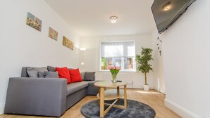 Una sala moderna con un sofá gris, una pequeña mesa redonda con un jarrón de flores, una planta en maceta y un televisor de pantalla plana montado en la pared.