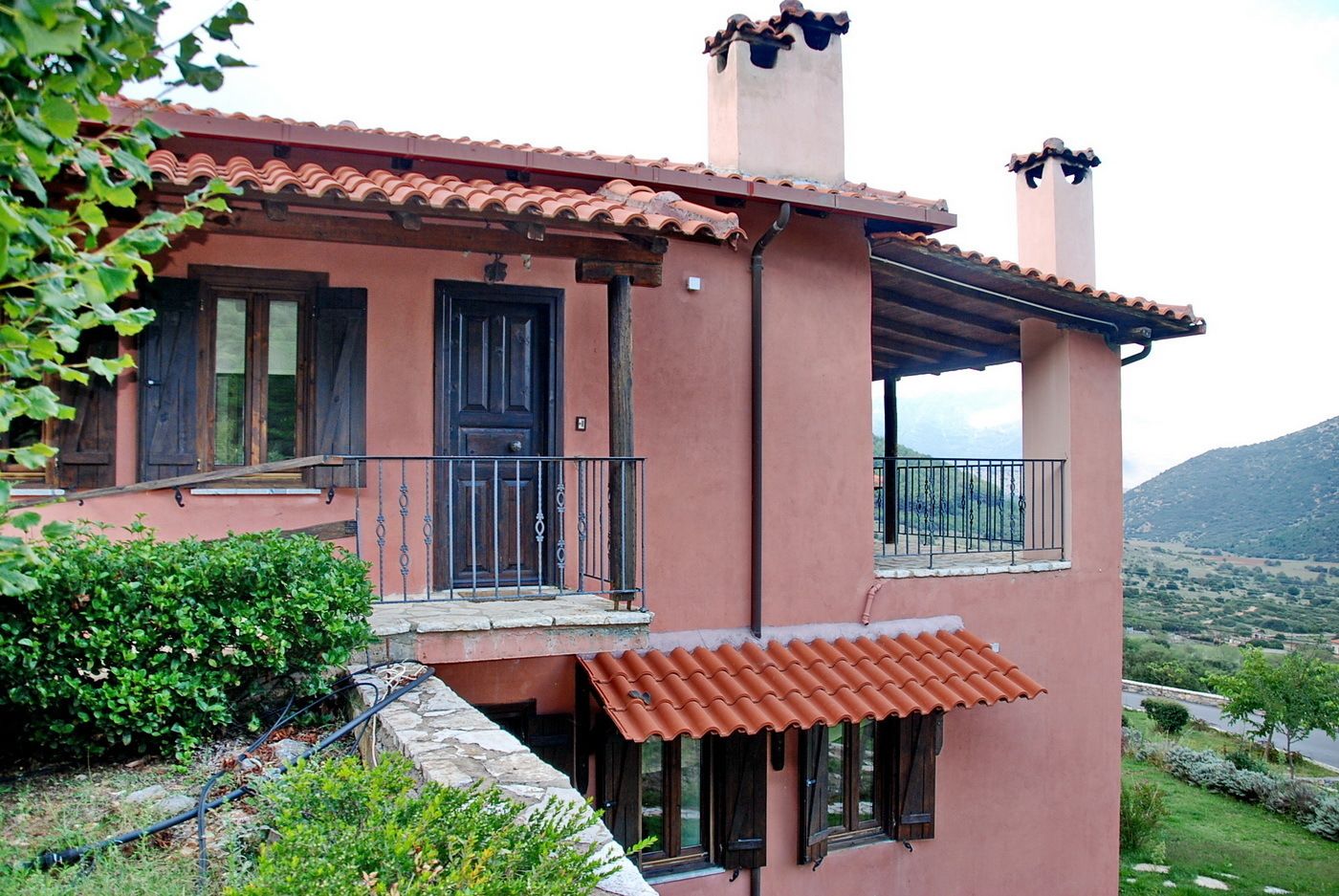 A two-story house with a red-tiled roof, a balcony with a metal railing, and a stone wall in front.