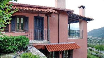 A two-story house with a red-tiled roof, a balcony with a metal railing, and a stone wall in front.