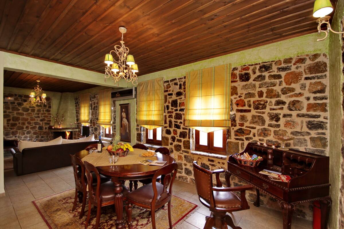A dining area with a wooden table, chairs, and a chandelier. There is a stone wall and a sofa in the background.