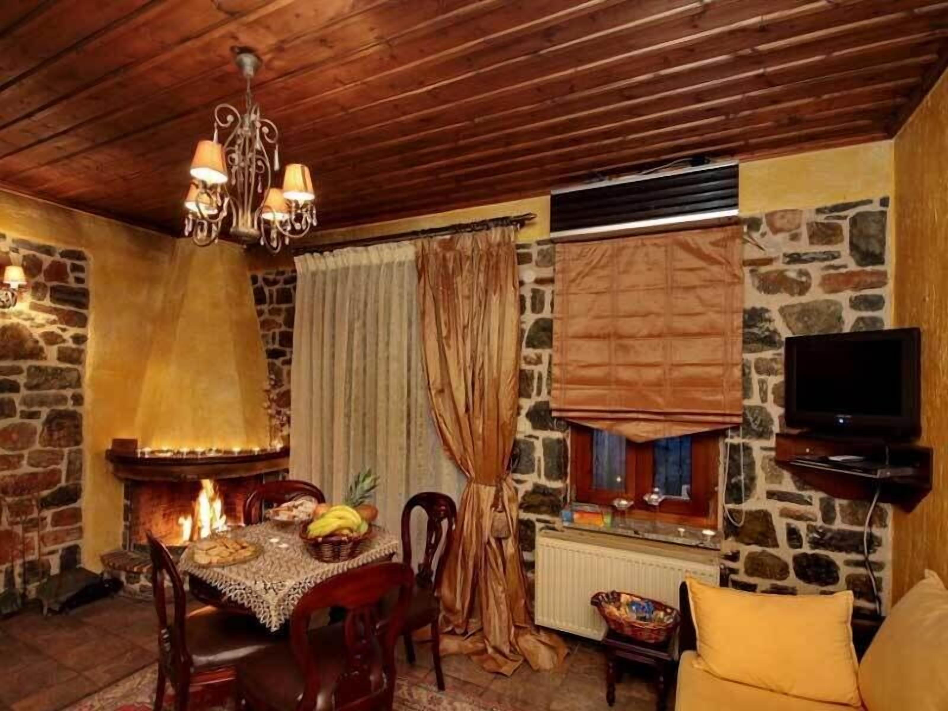 A cozy room with a fireplace, wooden ceiling, stone walls, a dining table with chairs, a television, and a window with curtains.