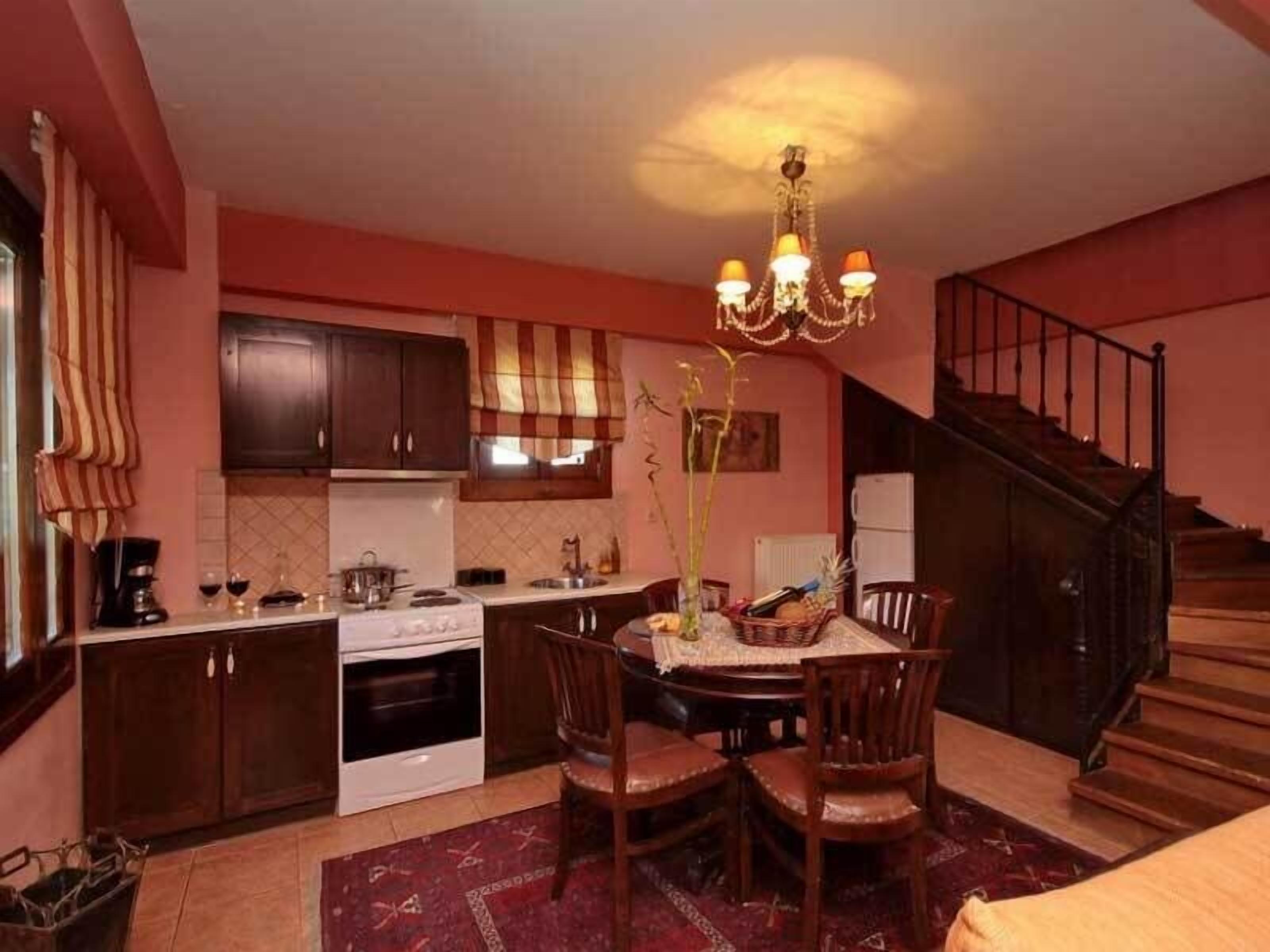 A compact kitchen with dark wooden cabinets, a white stove, and a dining area with a table and chairs. A chandelier hangs above the dining table, and a staircase is visible to the right.