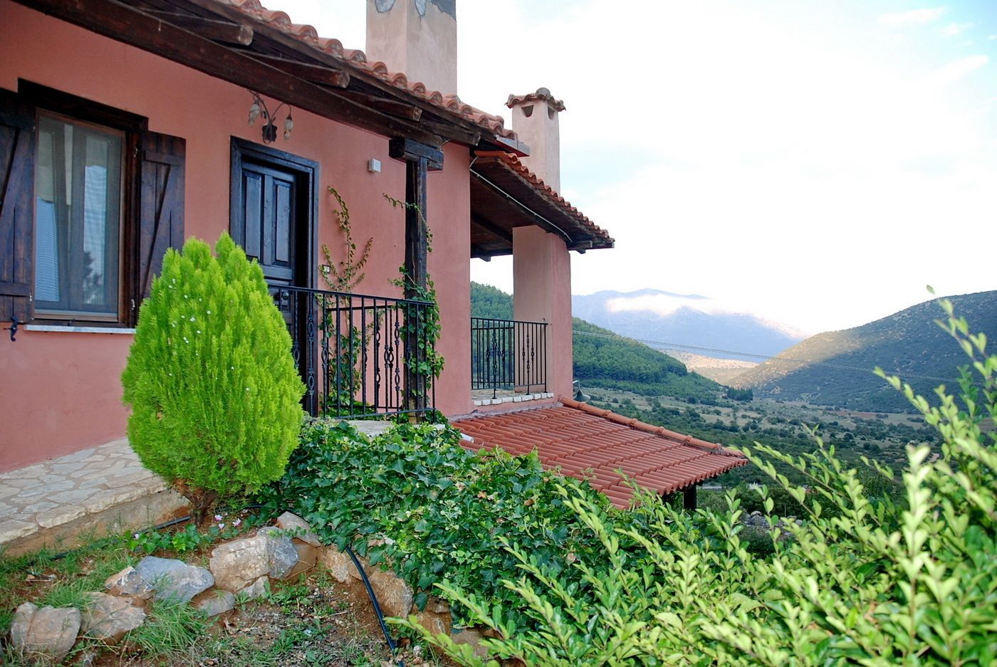 A house with a red-tiled roof, a balcony with a black railing, and a view of a valley with mountains in the background.