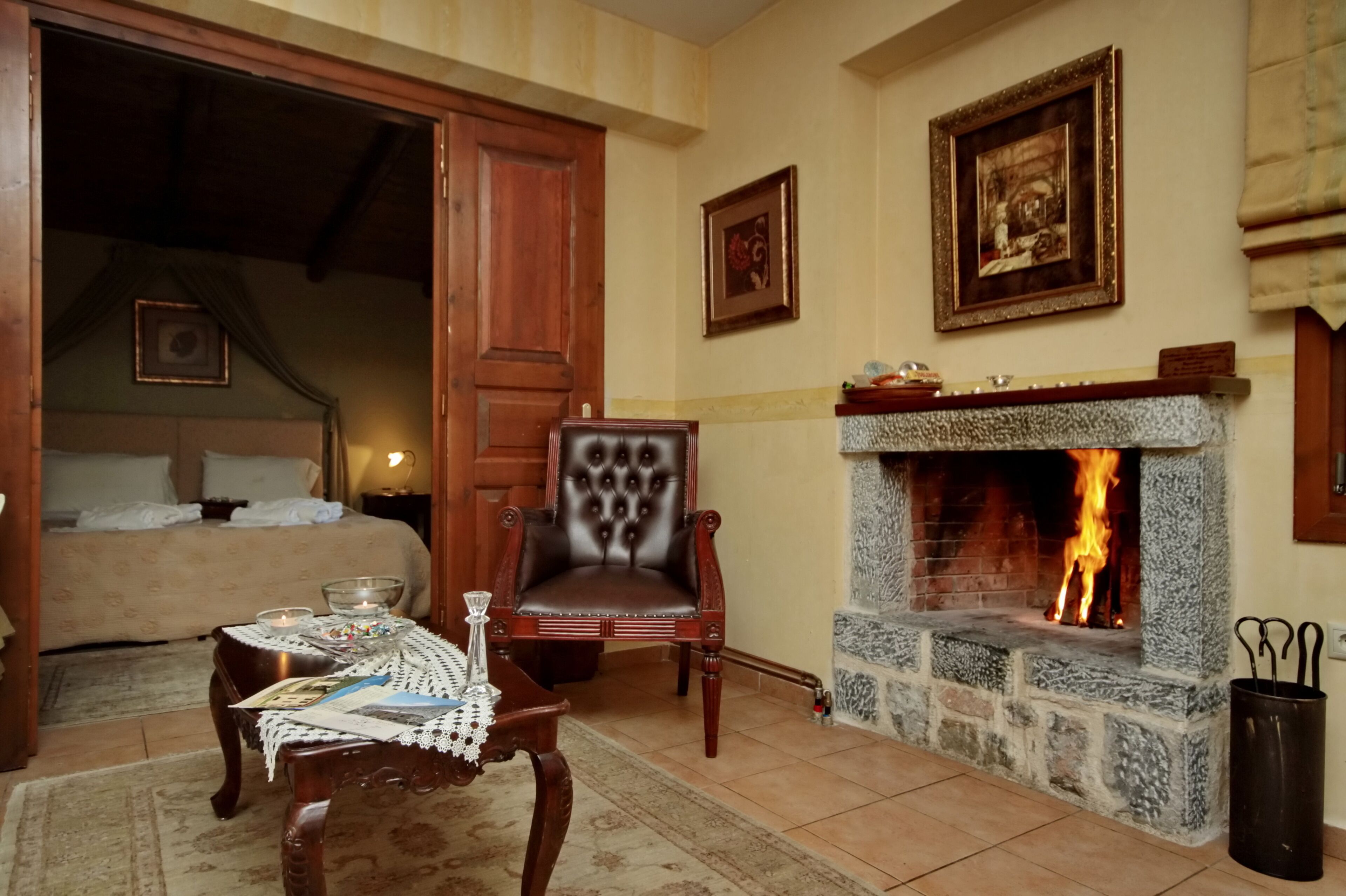 A cozy room with a stone fireplace, a wooden chair, a table with a lace tablecloth, and a bed with a headboard.