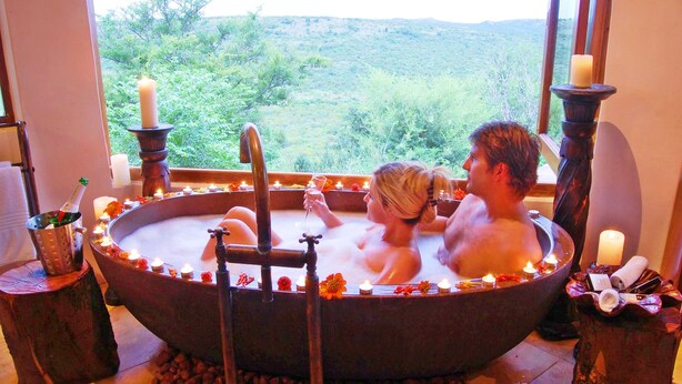 A couple in a large, round bathtub with candles and flowers around the edges, set against a scenic view. - Umzolozolo Private Safari Lodge (Ladysmith)