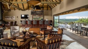 A spacious living area with a large TV, bar, and seating. A balcony overlooks a scenic view. - Umzolozolo Private Safari Lodge (Ladysmith)