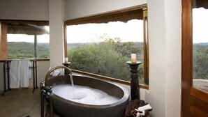 A bathroom with a large bathtub, a window with a view of a forest, and a towel rack with white towels. - Umzolozolo Private Safari Lodge (Ladysmith)