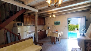A rustic kitchen with wooden cabinets, a dining area with a table and chairs, a sofa, and a view of a swimming pool. - Villa Sofas (Platanias)