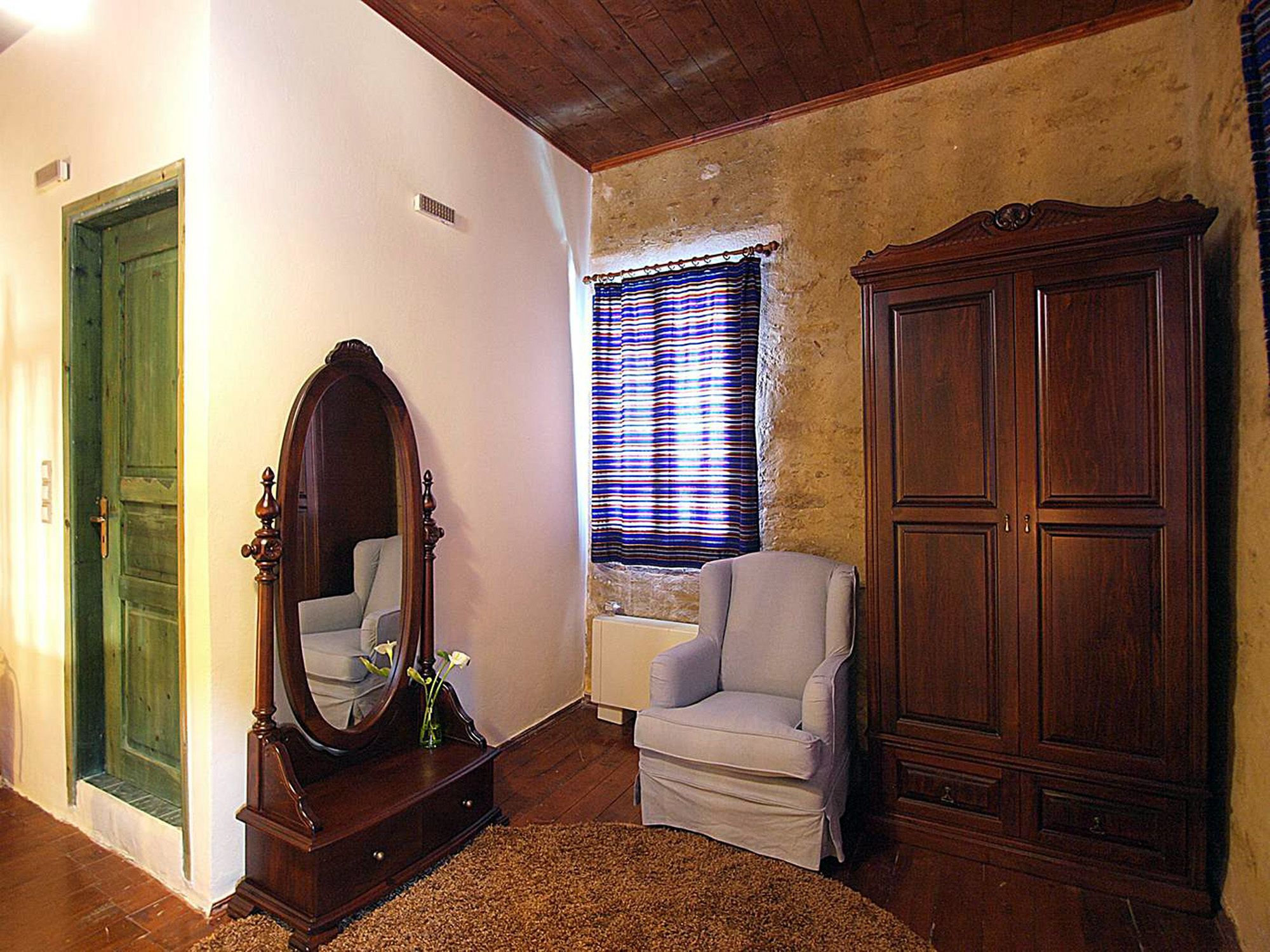 Une pièce avec un plancher en bois, une porte verte, un miroir, une chaise blanche, une fenêtre avec des rideaux à rayures et une grande armoire en bois.