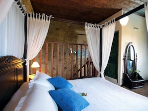 Een slaapkamer met een bed, witte gordijnen, een houten hoofdbord, een nachtkastje, een lamp, een spiegel en uitzicht op een balkon.