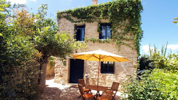 A rustic stone building with ivy-covered walls, a stone patio with a wooden table and chairs, and a yellow umbrella providing shade. - Villa Sofas (Platanias)
