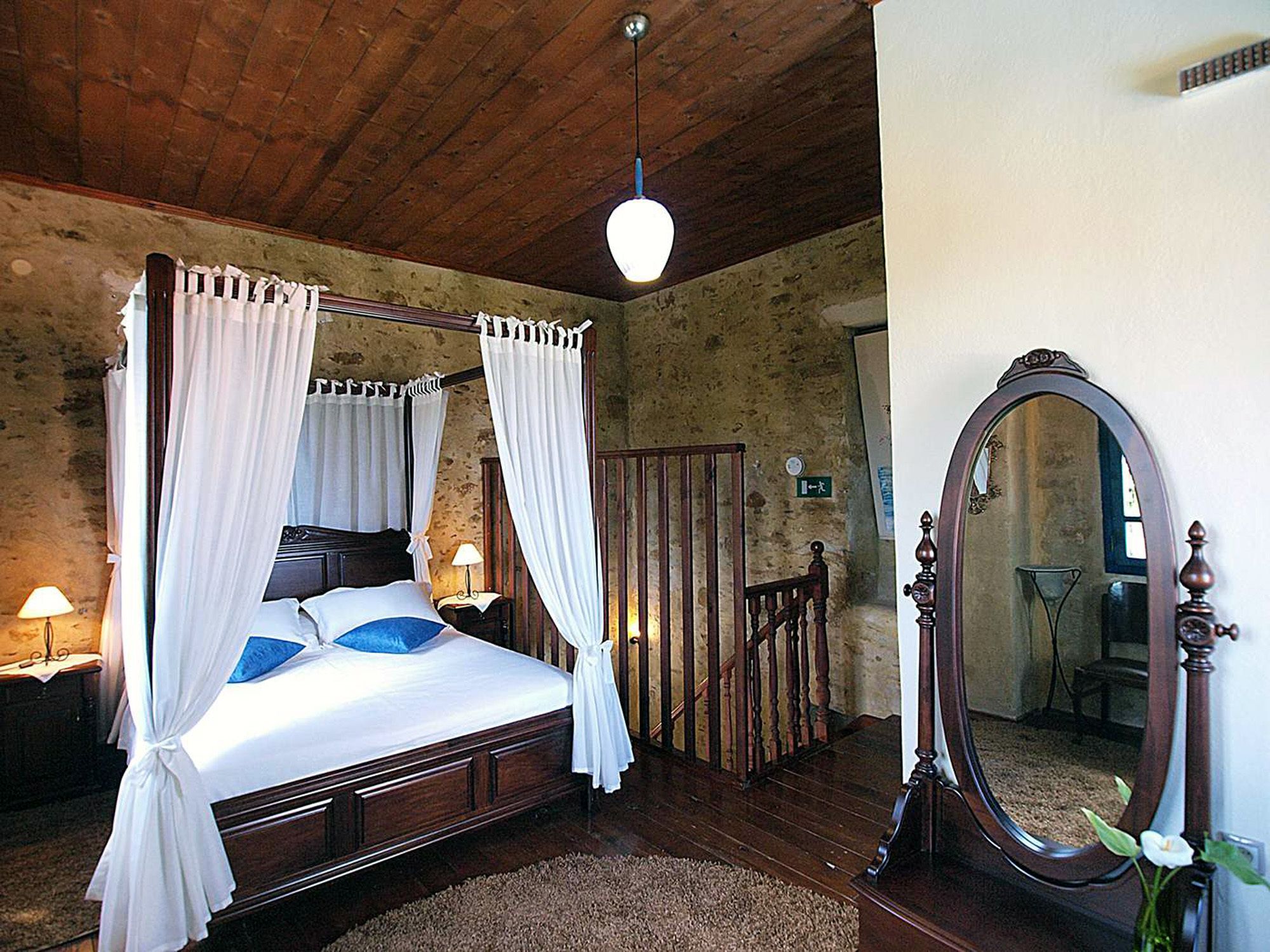 A four-poster bed with white bedding and blue pillows, a wooden ceiling, a wooden floor, a mirror, and a small table with a lamp.